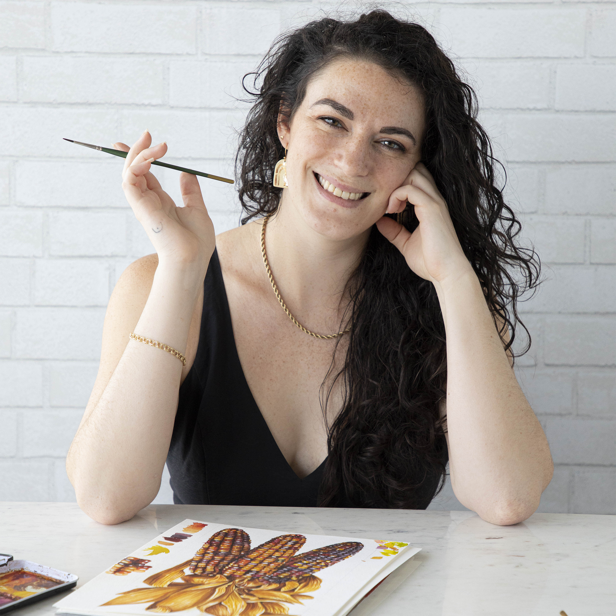 Christie, an illustrator at Hester & Cook, smiling with a paint brush in her hand and her artwork in front.