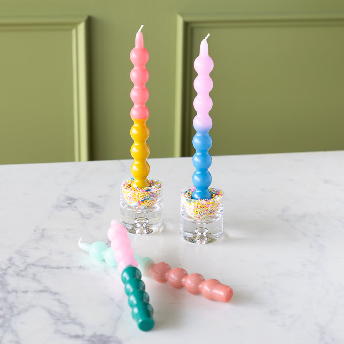 Four Colorful Bubble Taper Candles in pastel hues, two upright in sprinkle-filled glass holders and two lying on a white marble surface.