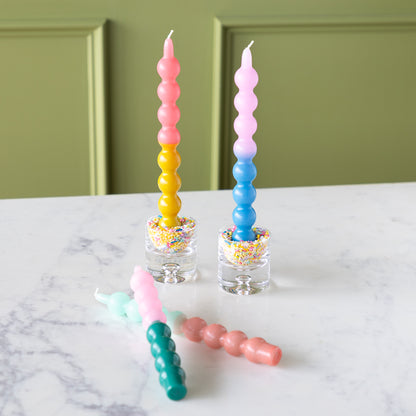 Four Colorful Bubble Taper Candles in pastel hues, two upright in sprinkle-filled glass holders and two lying on a white marble surface.