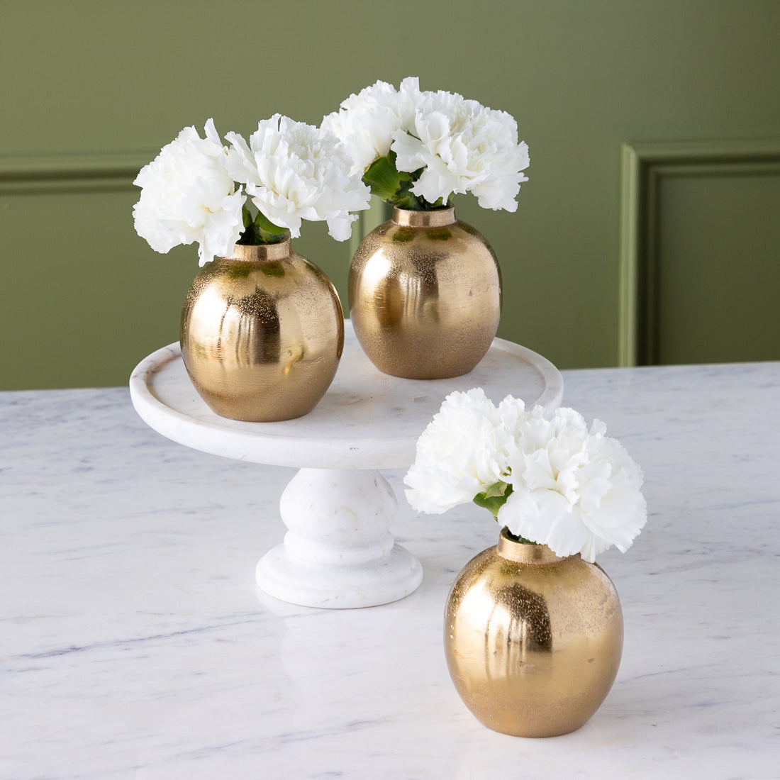 Three round Gold Bud Vases with white flowers are arranged on a white marble cake stand and table against a green wall background.