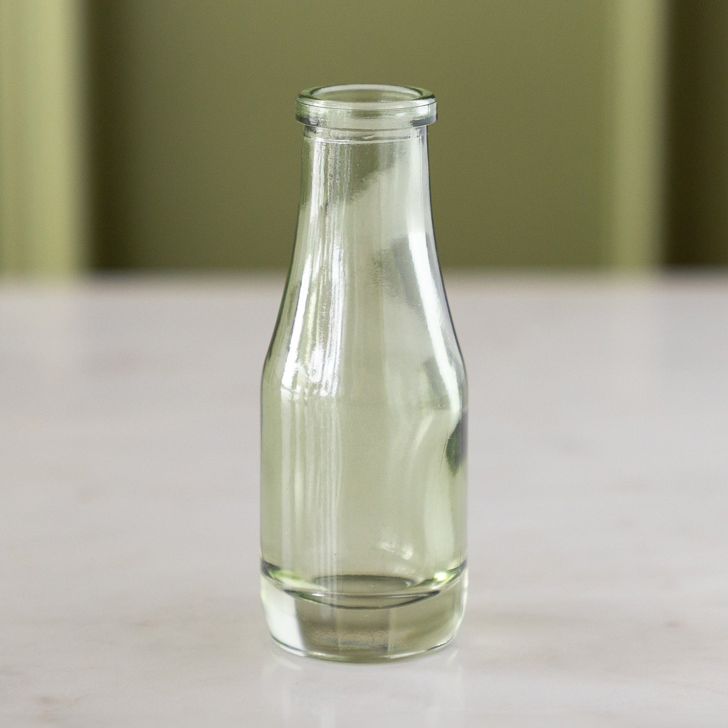 A small, empty, clear green glass bottle, reminiscent of vintage bud vases, stands on a marble table against a light green background.