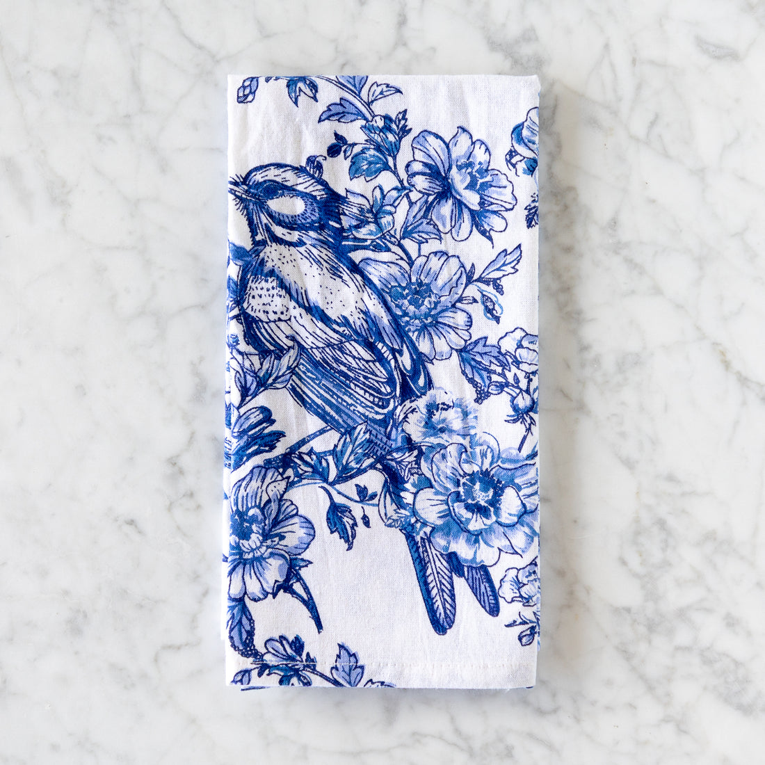 A white cotton napkin, made in India, features a blue illustration of a bird perched among flowers and is displayed on a marble table.