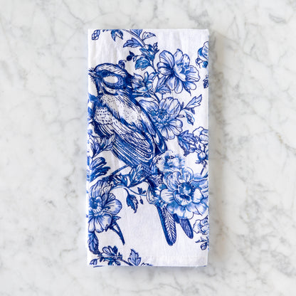 A white cotton napkin, made in India, features a blue illustration of a bird perched among flowers and is displayed on a marble table.