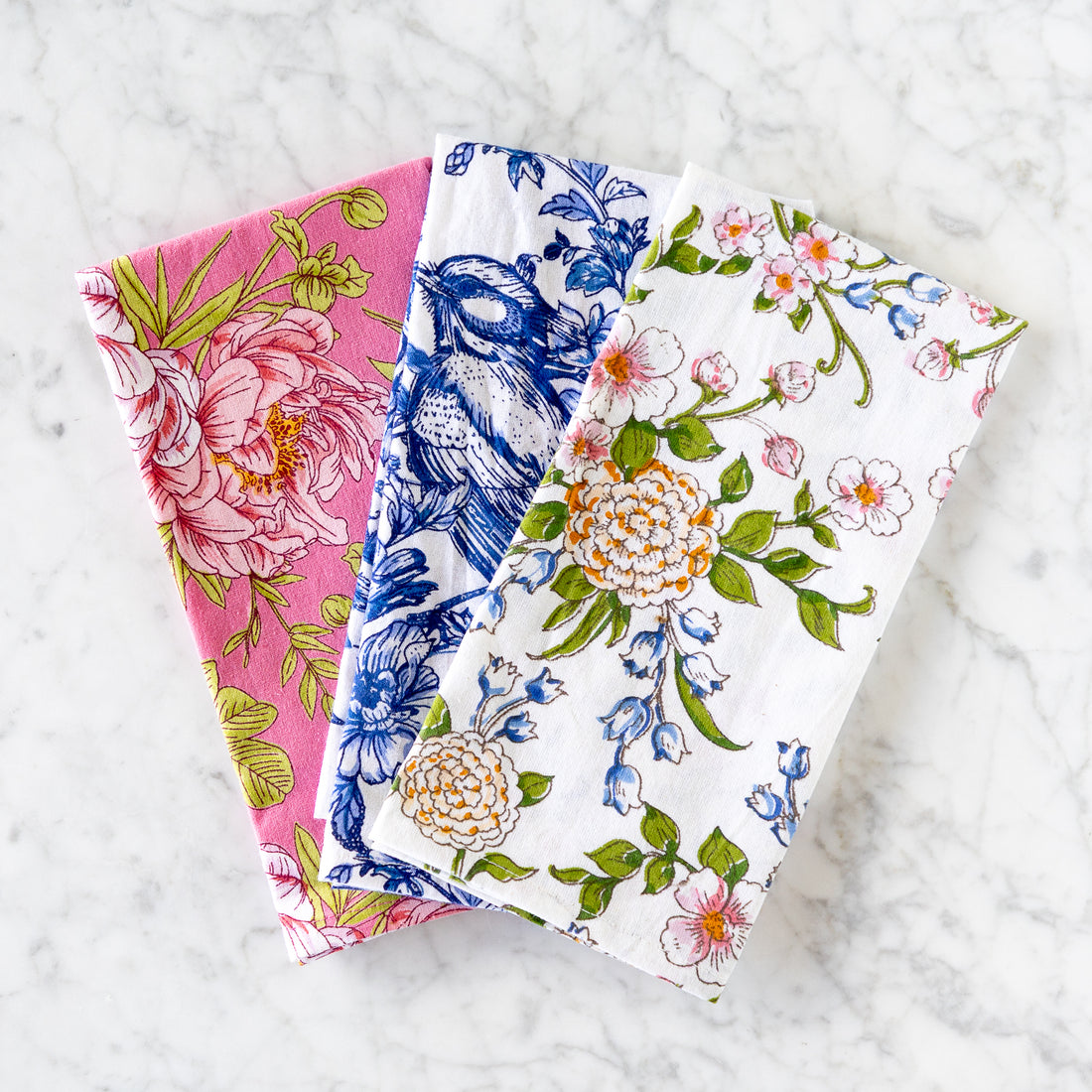 Three folded cotton napkins, Eloise Rose, Bluebird Floral and White Floral, are arranged on a marble surface. 