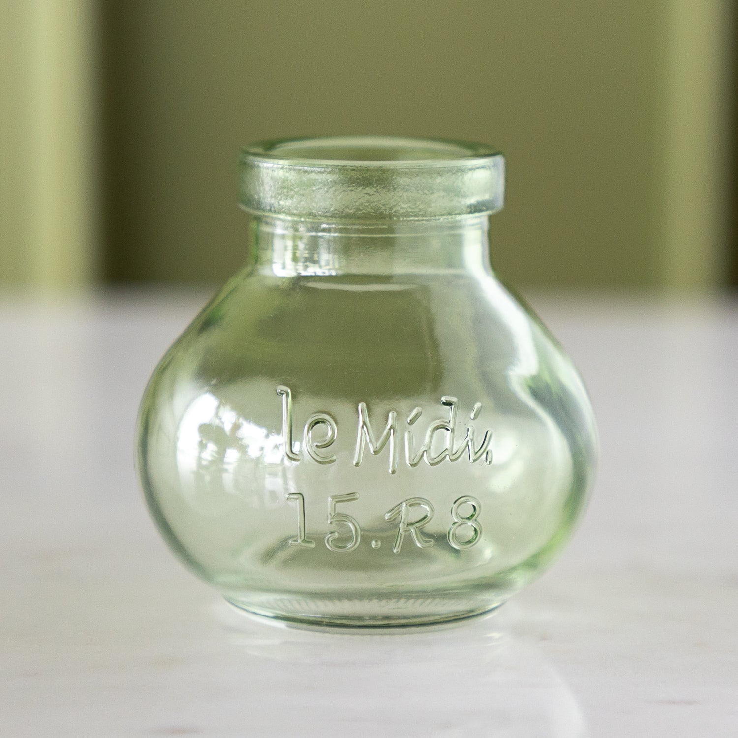 A small, empty, clear green glass bottle with &quot;le Midi. 15.R8&quot; embossed on the front, reminiscent of vintage bud vases, stands on a marble table against a light green background.