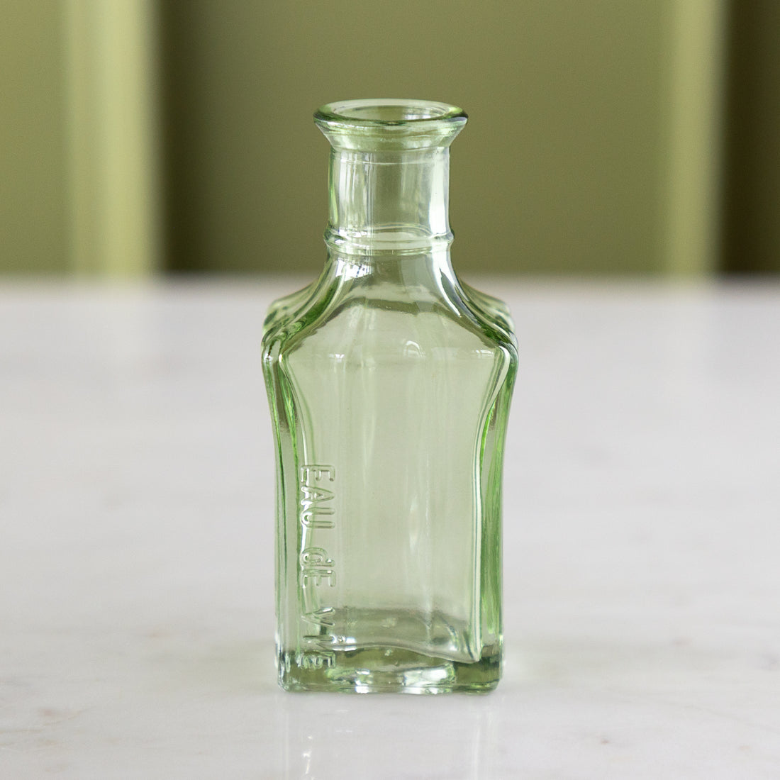 A small, empty, clear green glass bottle with &quot;Eau de Vie&quot; embossed on the side, reminiscent of vintage bud vases, stands on a marble table against a light green background.