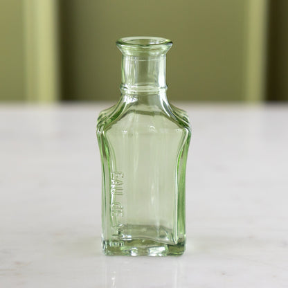 A small, empty, clear green glass bottle with &quot;Eau de Vie&quot; embossed on the side, reminiscent of vintage bud vases, stands on a marble table against a light green background.
