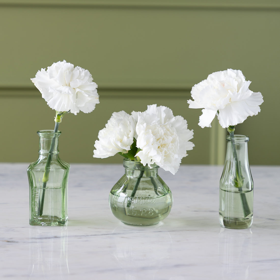 Three Green Glass Bud Vases with white flowers in them are displayed on a marble table in a row.