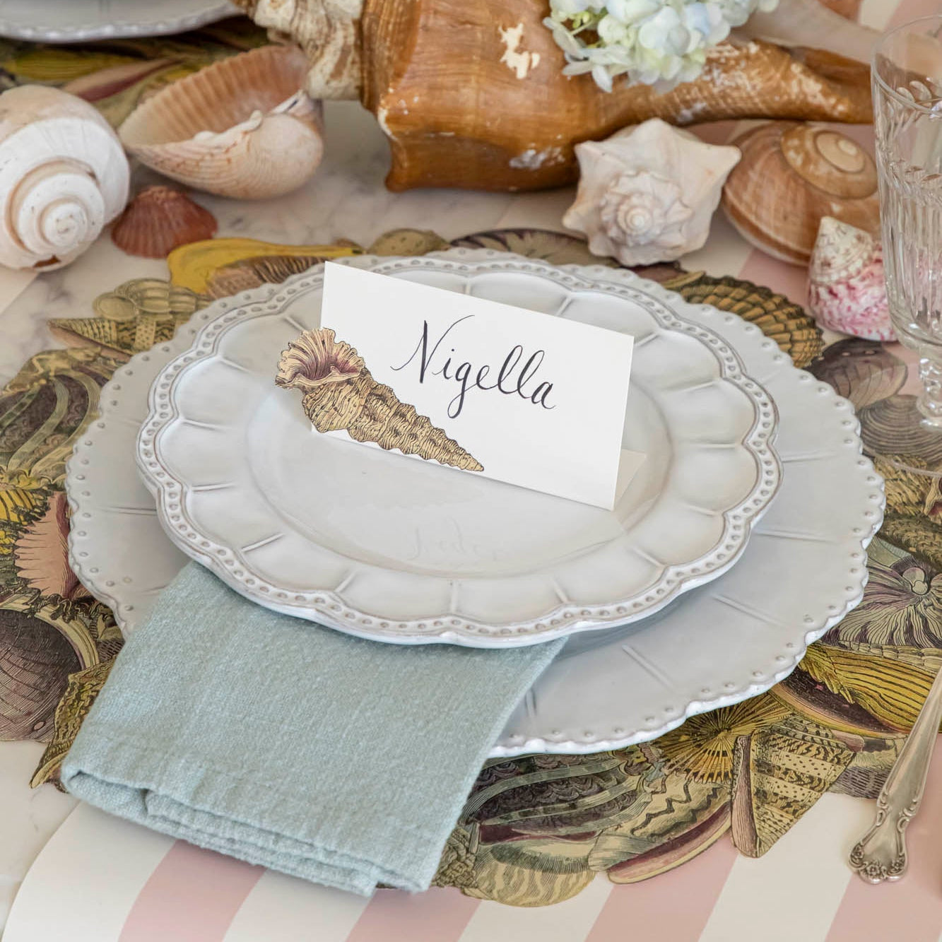 Shell Place Card with &quot;Nigella&quot; written in the white space, on a set of dinner plates on an elegant place setting.