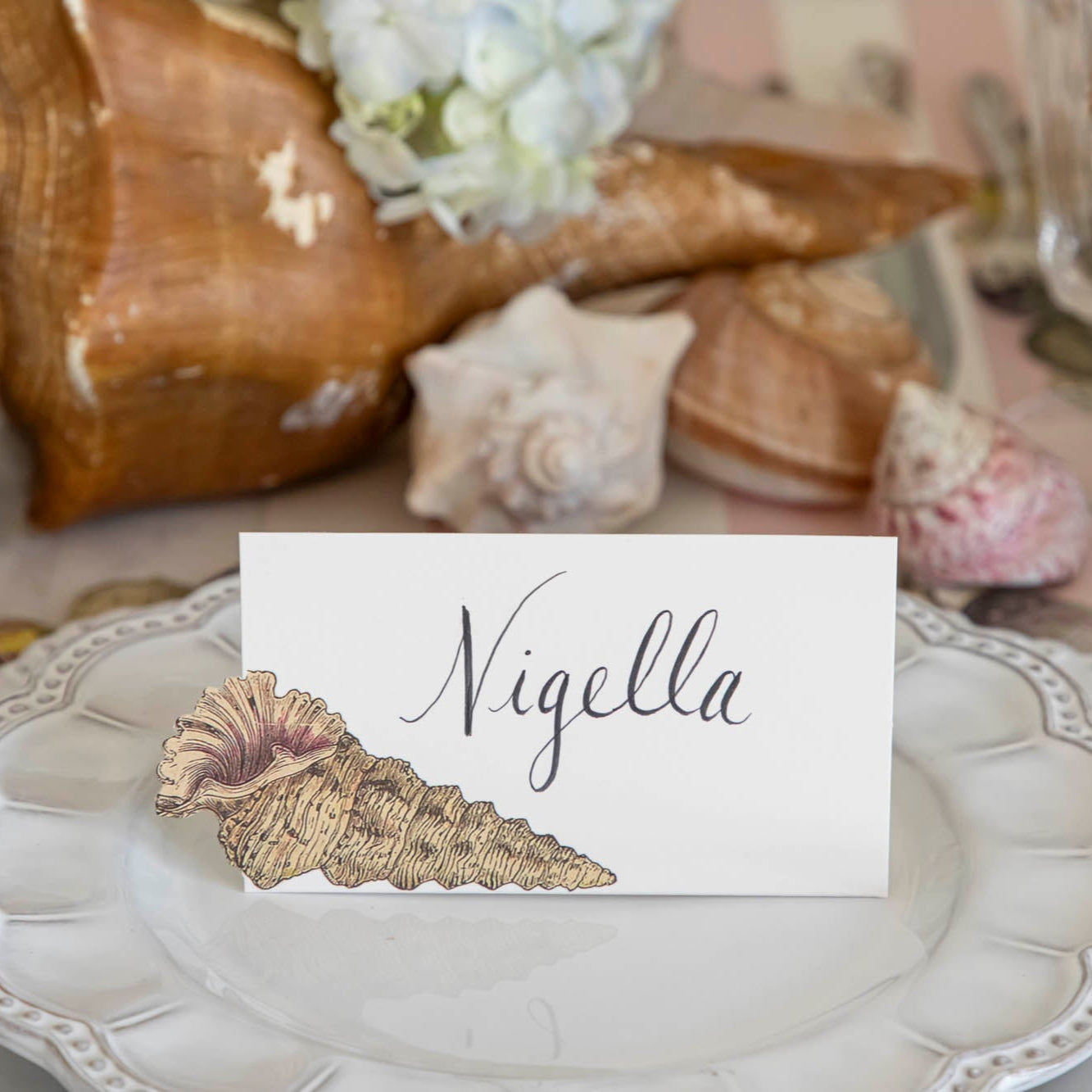 Shell Place Card with &quot;Nigella&quot; written in the white space, on a set of dinner plates.