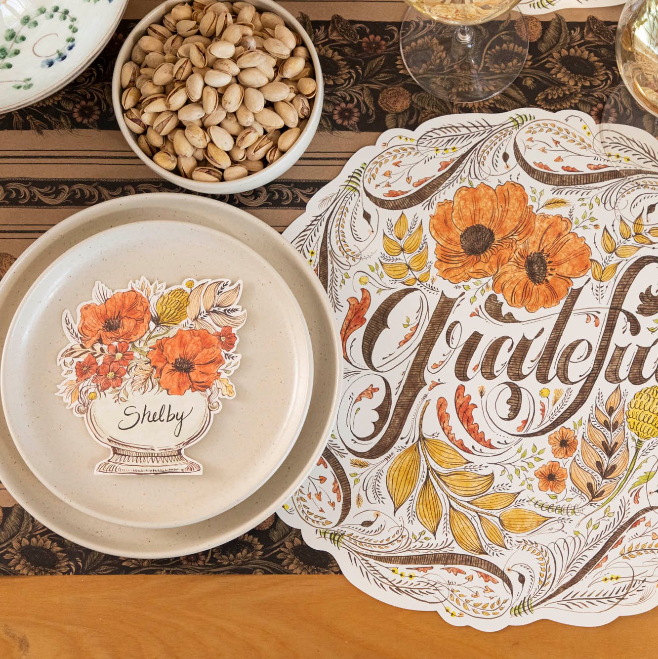 The Autumn Arrangement Place Card with the name &quot;Shelby&quot; written on it, on a fall themed table setting with a floral placemat with &quot;Grateful&quot; written on it.