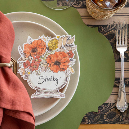 The Autumn Arrangement Place card with the name &quot;Shelby&quot; written on it, on top of salt dinner plates, a Green French Frame placemat and Vintage Sunflower Stripe runner underneath.