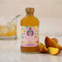 A bottle of Hester & Cook Peach Basil Nectar Syrup next to a glass of ice and a slice of peach garnished with basil.