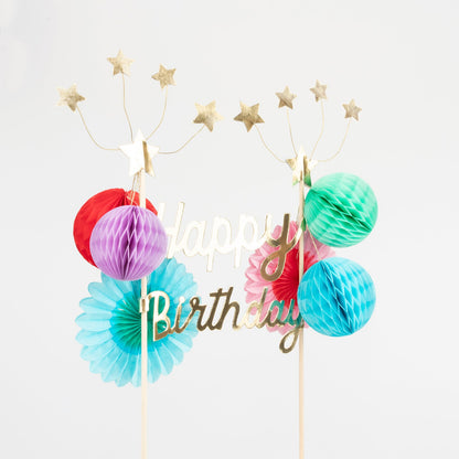 A Happy Birthday Honeycomb Cake Topper with gold foil stars and pom poms by Meri Meri.