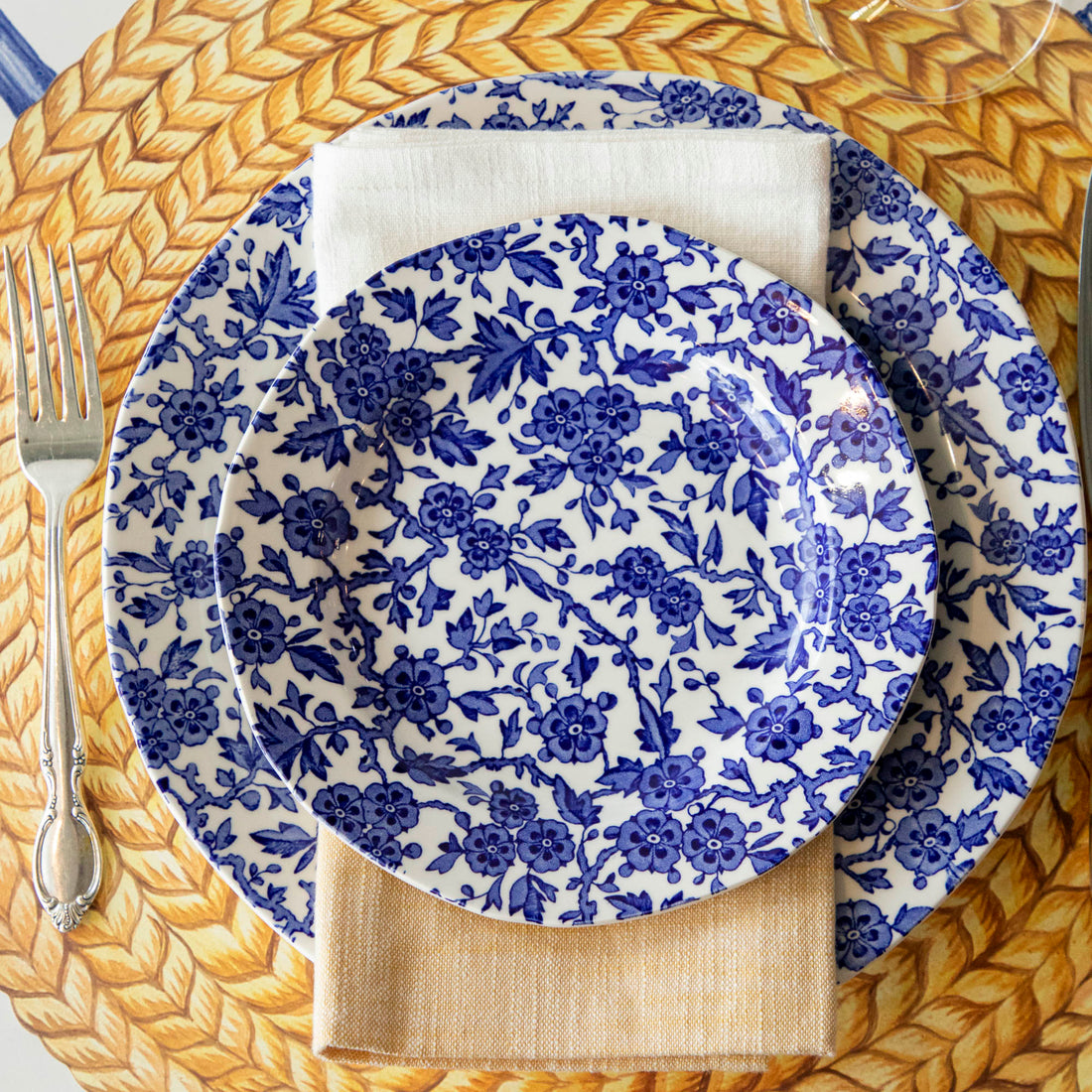 A Burleigh Blue Arden dinnerware plate on a wicker placemat with a folded napkin and a fork to the side, also noted as dishwasher safe.