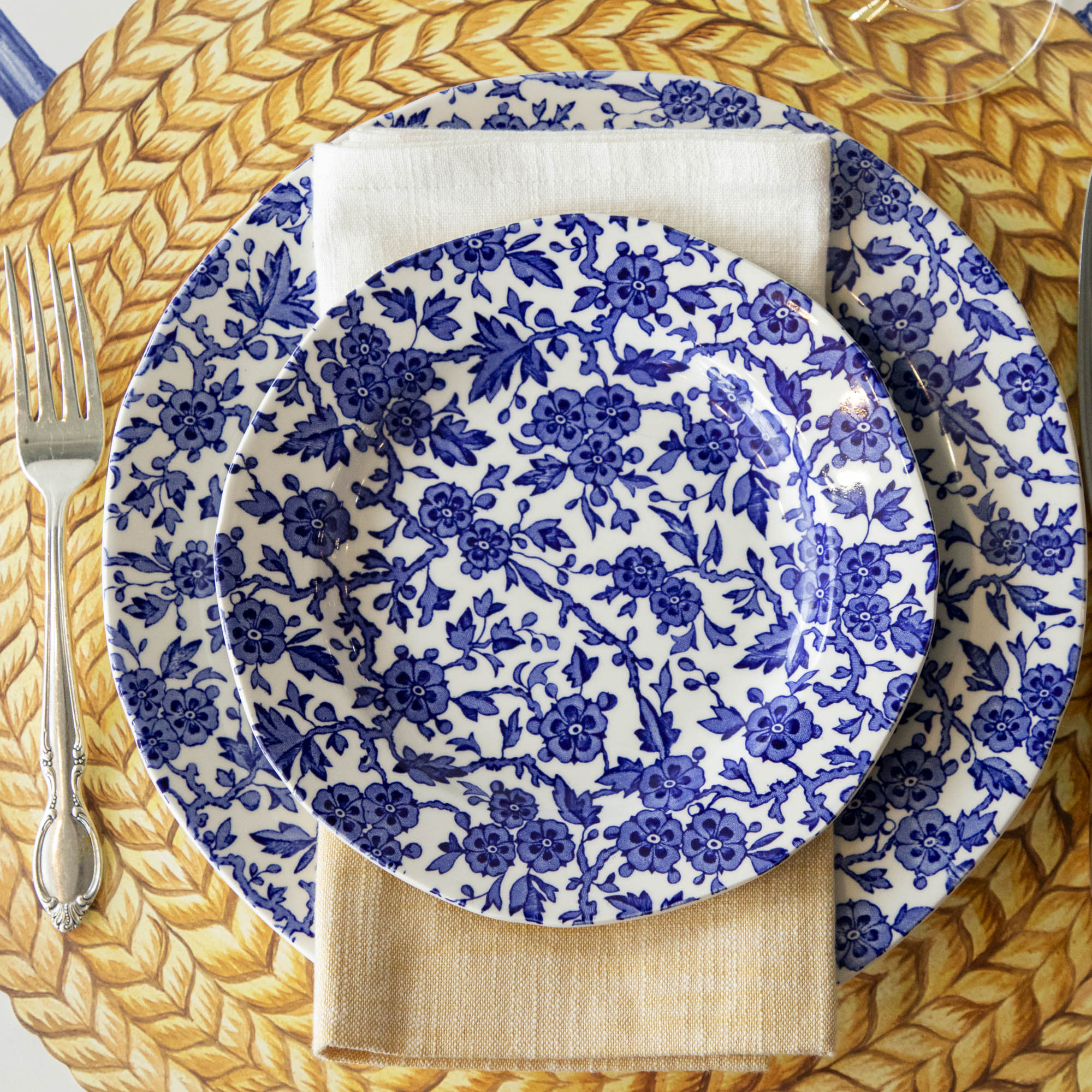 A Burleigh Blue Arden dinnerware plate on a wicker placemat with a folded napkin and a fork to the side, also noted as dishwasher safe.