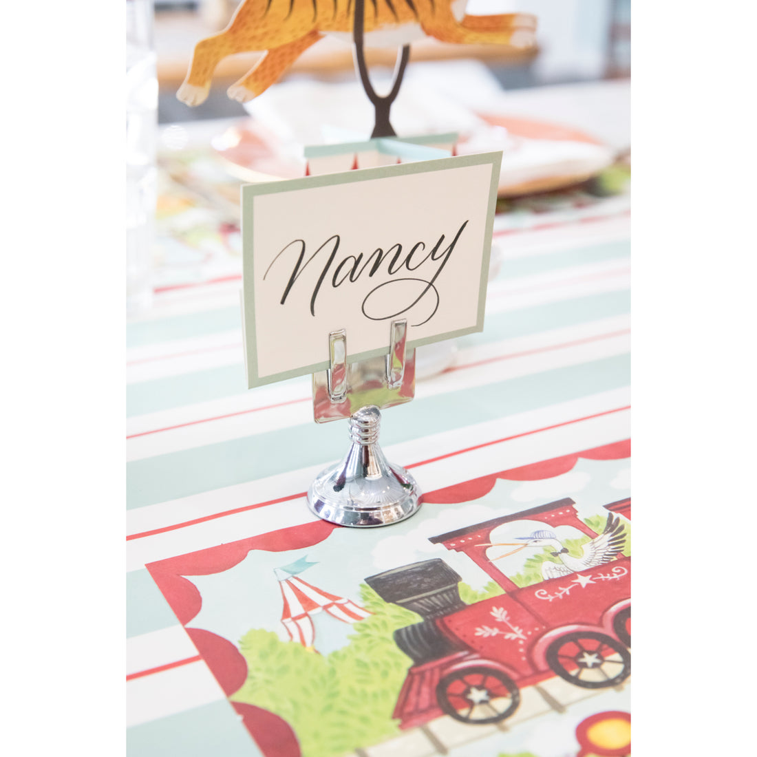 An elegant, chrome, short round stand topped by a flat card-holder with two flat prongs, holding a place card reading &quot;Gwen&quot;.