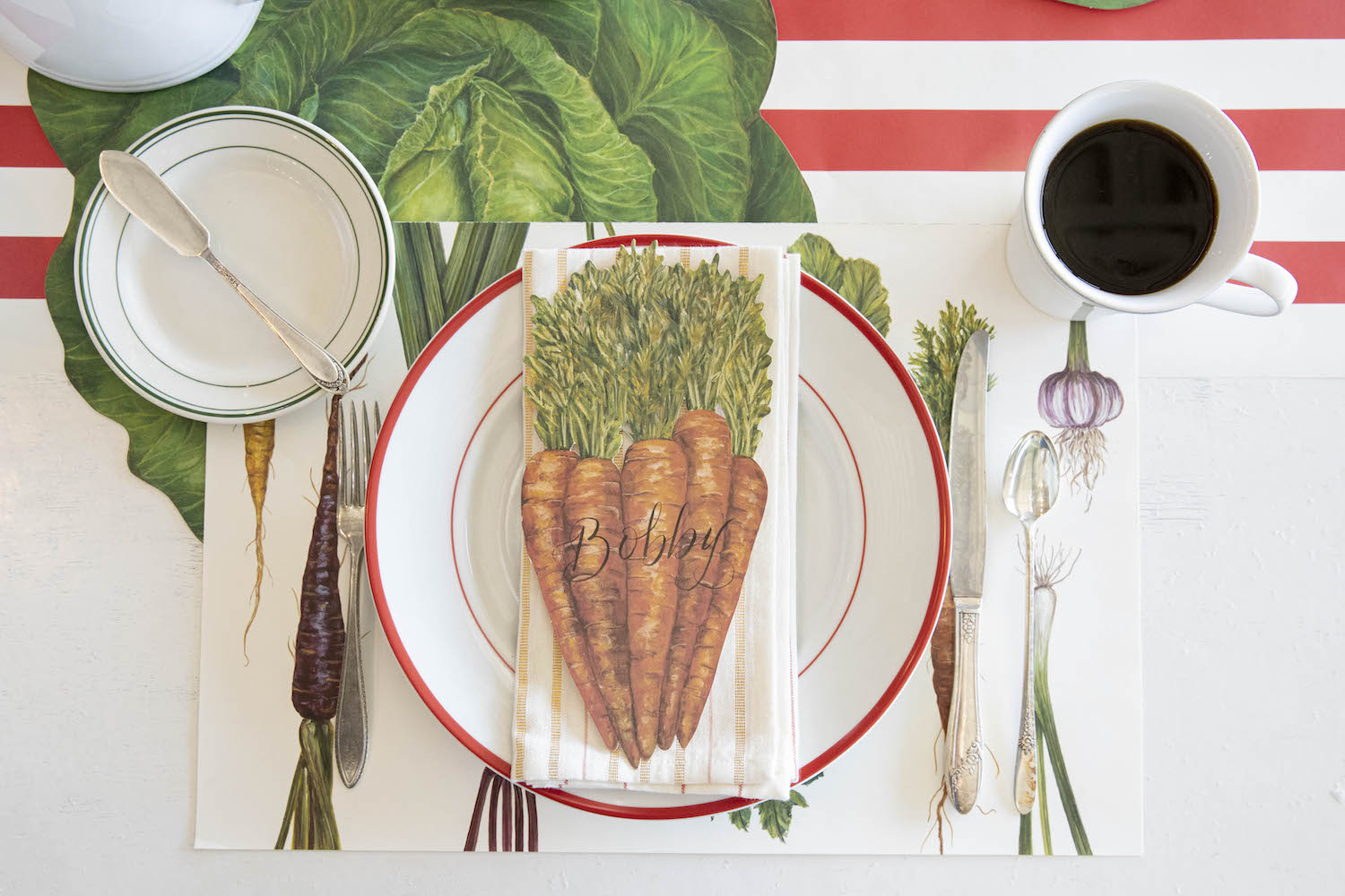 A Carrots Table Accent labeled &quot;Bobby&quot; resting on the plate of a vegetable-themed place setting, from above.