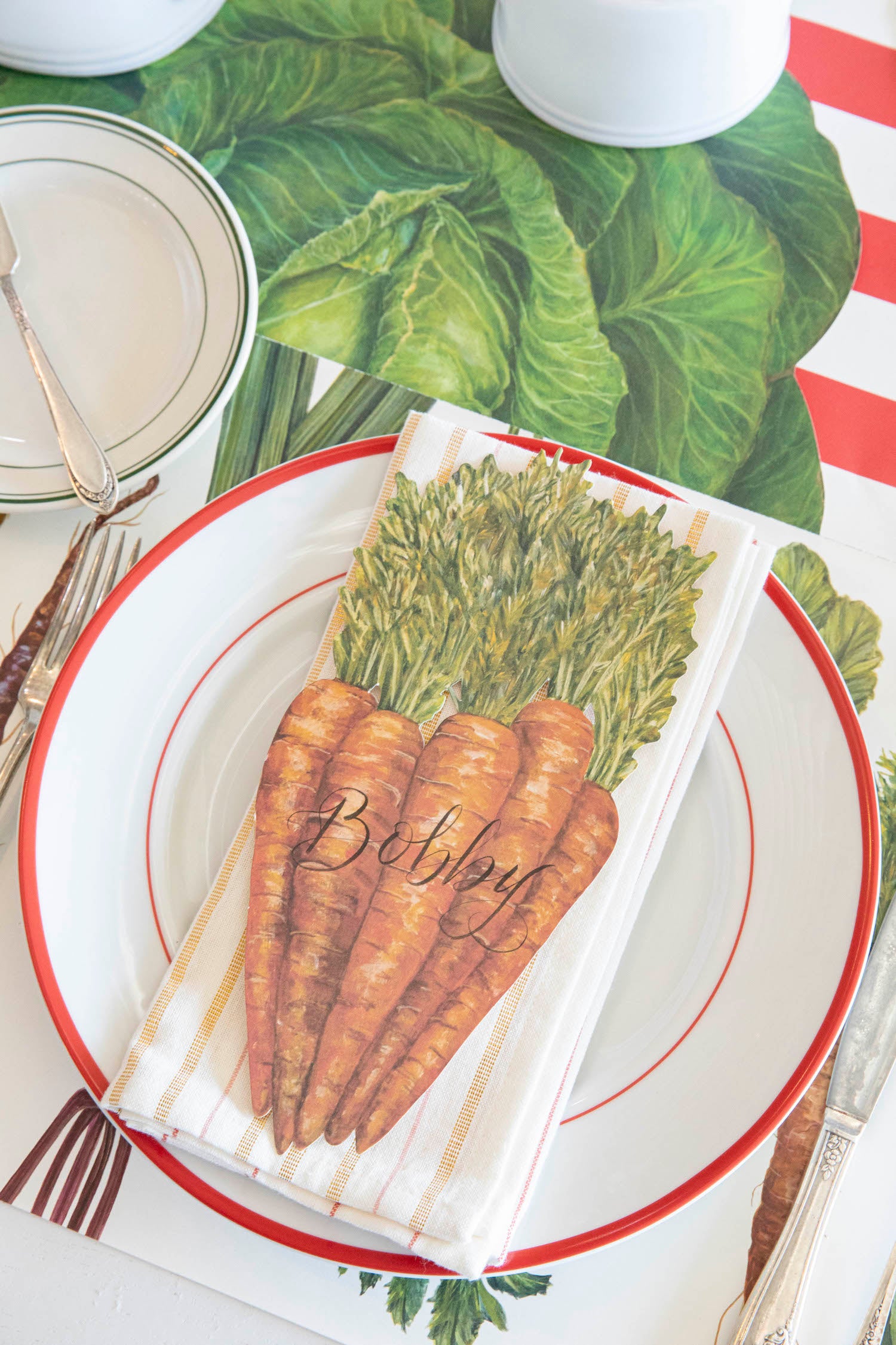 A Carrots Table Accent labeled &quot;Bobby&quot; resting on the plate of a vegetable-themed place setting, from above.