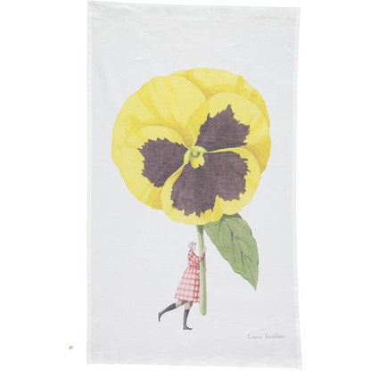A white tea towel featuring a whimsical illustration of a small woman holding a gigantic yelllow pansy by the stem. Art by Laura Stoddart. 