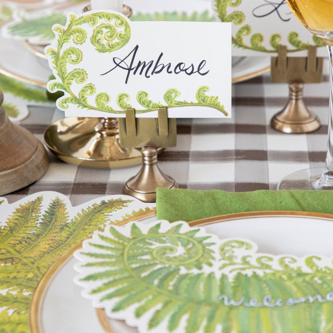 An elegant place setting featuring a Fiddlehead Fern Place Card labeled &quot;Ambrose&quot; standing in a brass place card holder.
