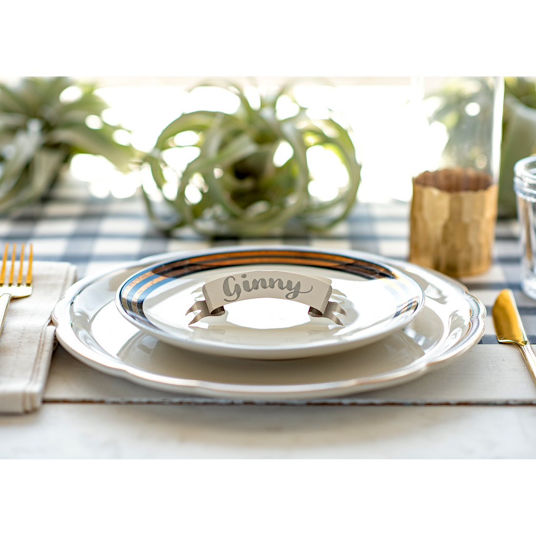 An elegant place setting featuring a Banner Place Card reading &quot;Ginny&quot; standing on the plate.