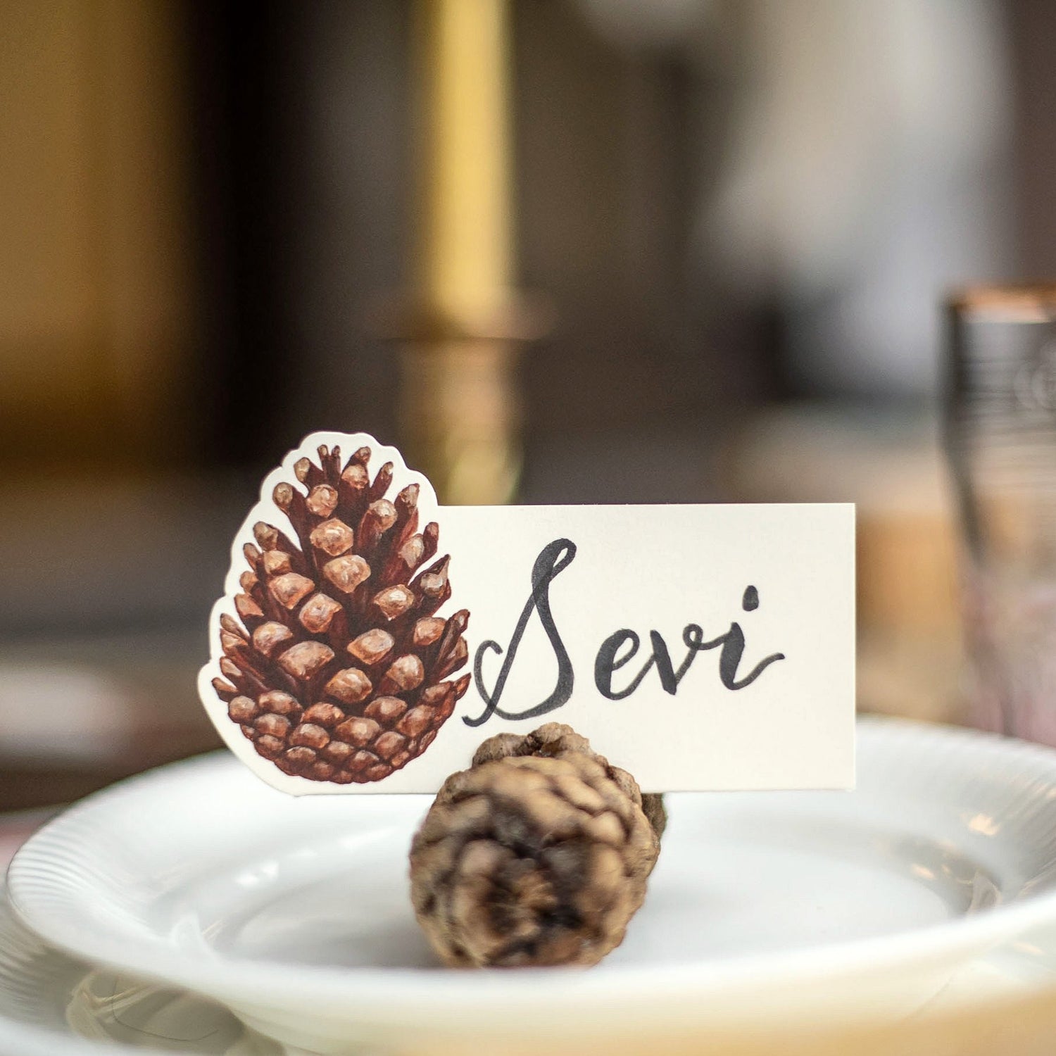 An elegant place setting featuring a Pinecone Place Card being held up by a real pinecone on a plate.