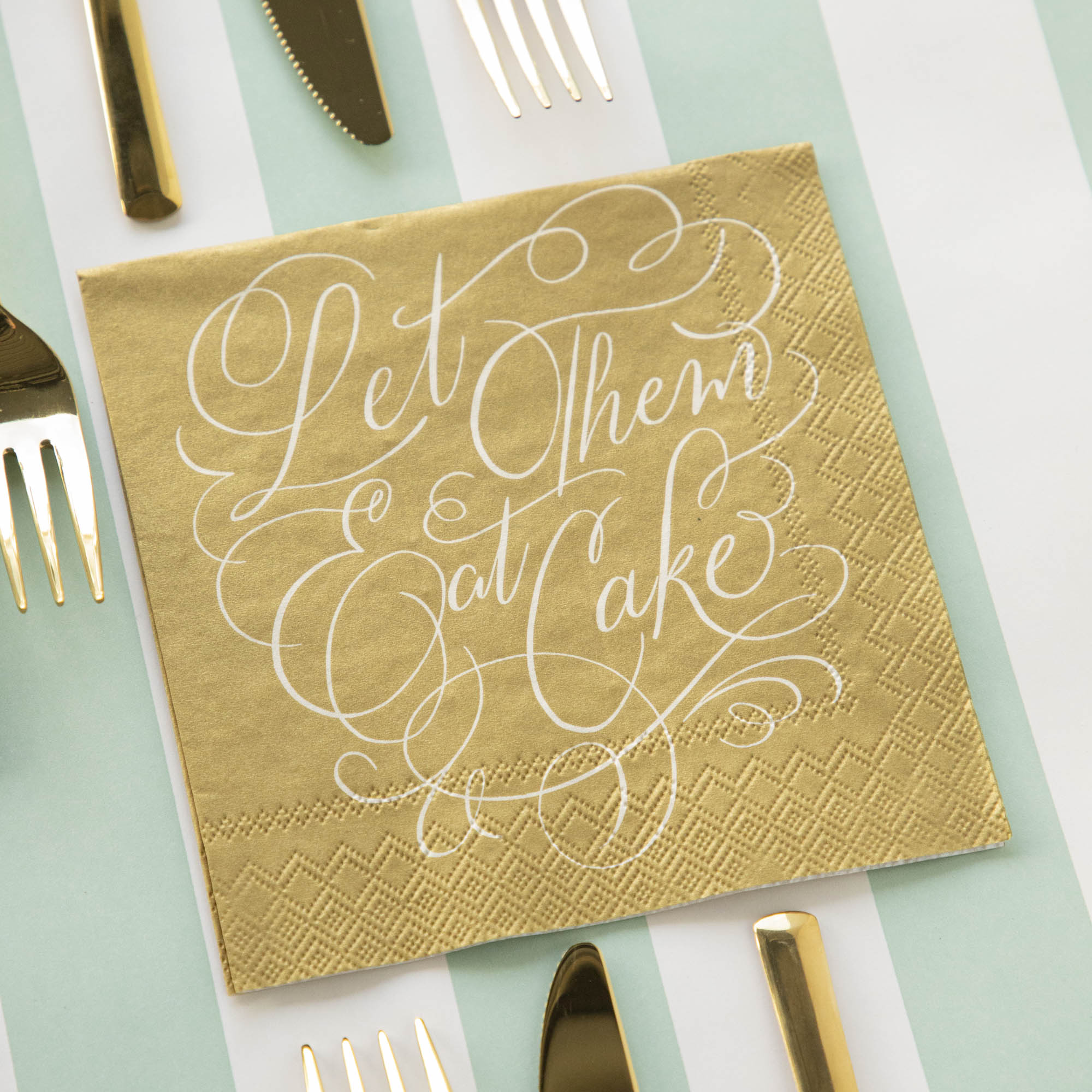 A Cake Cocktail Napkin on a striped table runner with gold cutlery.