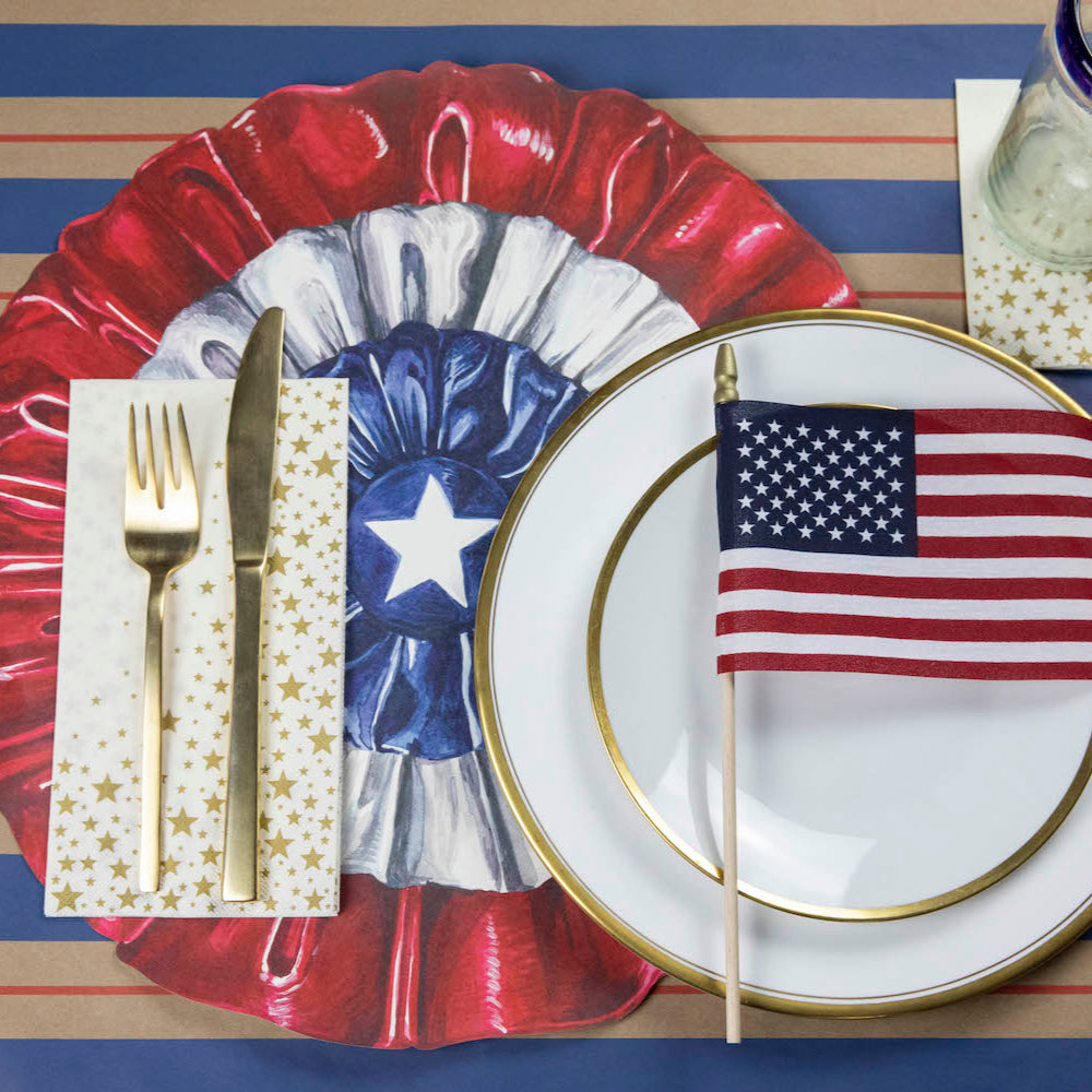 A patriotic place-setting with a Shining Star Guest Napkin with a gold fork and knife next to the plate, and a Shining Star Cocktail Napkin under the glass.