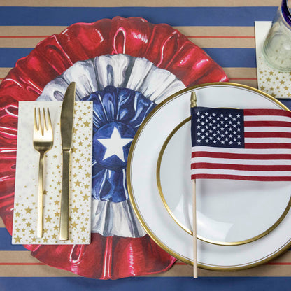 A patriotic place-setting with a Shining Star Guest Napkin with a gold fork and knife next to the plate, and a Shining Star Cocktail Napkin under the glass.