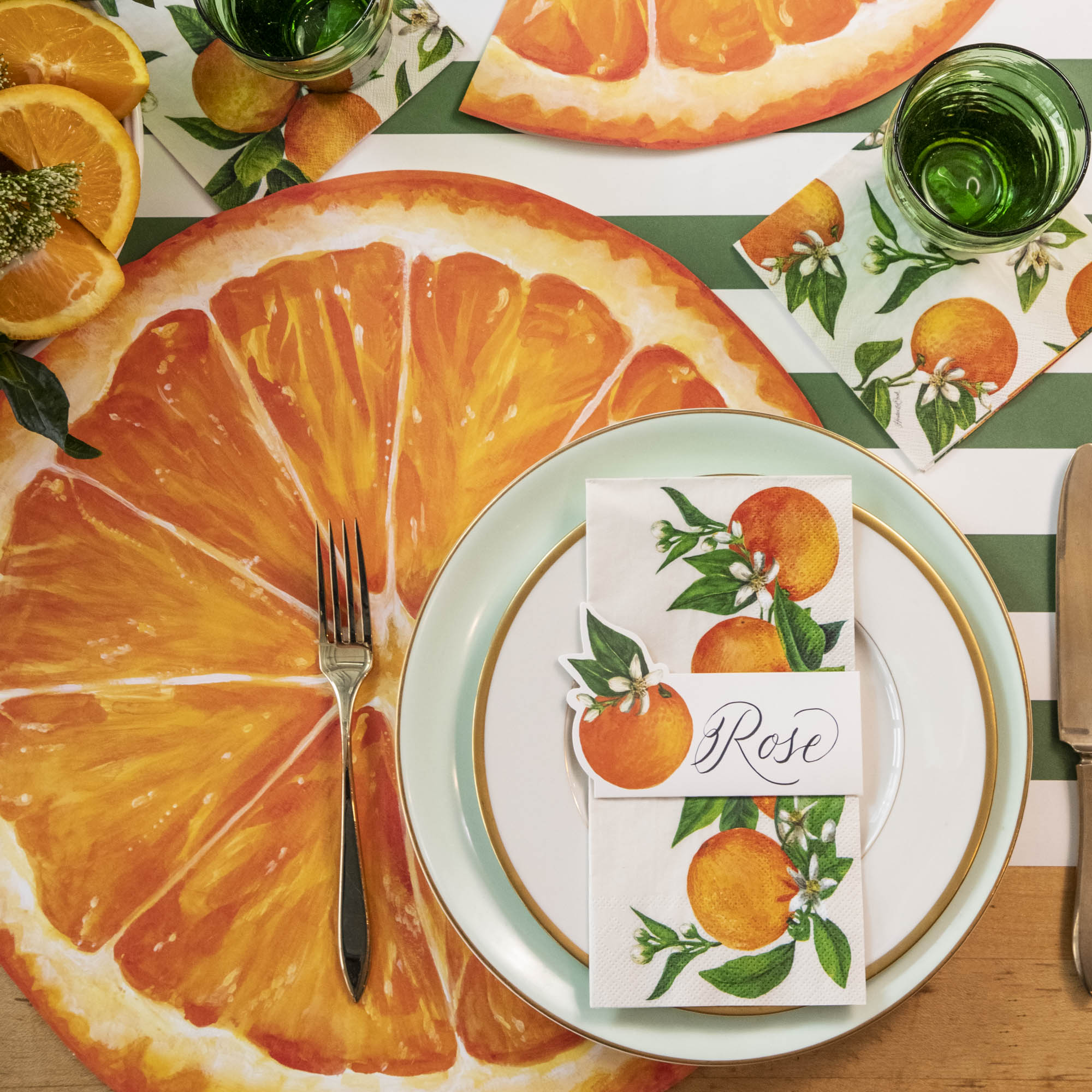 A vibrant citrus-themed place setting, featuring an Orange Orchard Guest Napkin centered on the plate, and an Orange Orchard Cocktail Napkin under the water glass.