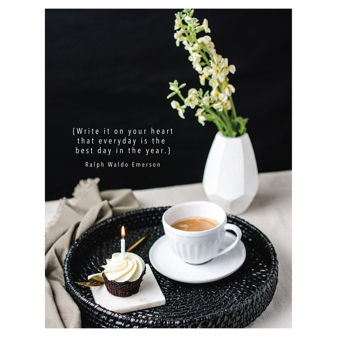 A photo of a round black tray with a cup of coffee and a cupcake with a lit candle, sitting on a white table with a white vase of flowers on a black background, with &quot;{Write it on your heart that everyday is the best day in the year.} Ralph Waldo Emerson&quot; printed in white.