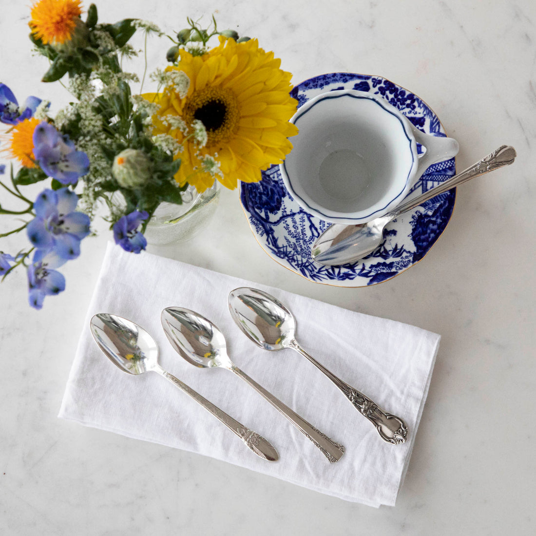 An elegant tea setting with an empty teacup and saucer, a Hester &amp; Cook Vintage Silver-Plate Tea Spoon Set of Four, and a bouquet of fresh flowers on a marble surface.