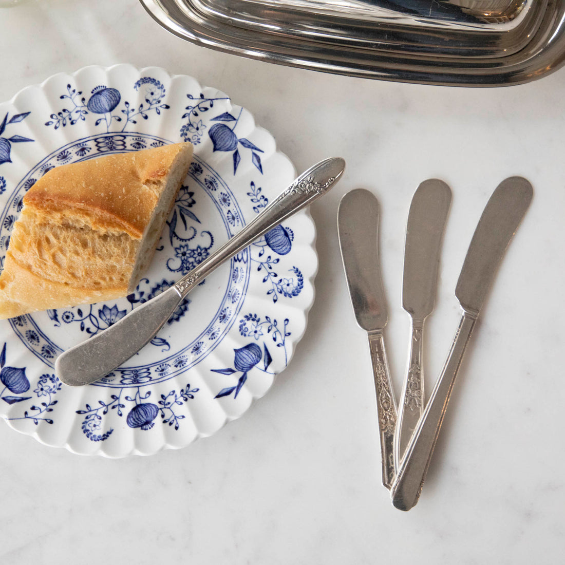 A piece of bread on a decorative plate beside a Hester &amp; Cook Vintage Silver-Plate Butter Spreader Set of Four on a marble surface.