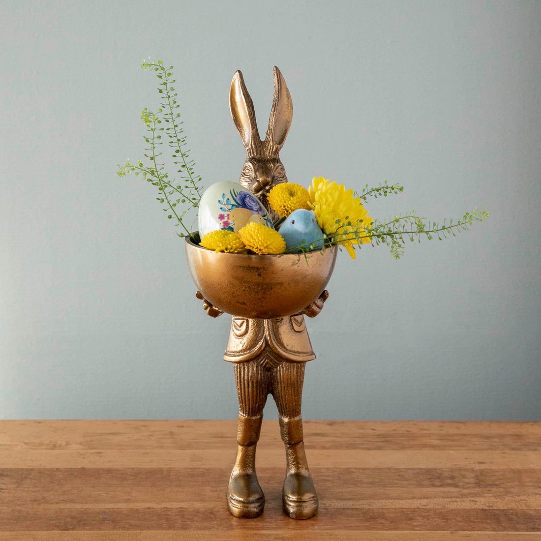 A well-dressed Hare Dish Stand by Accent Decor holding a round dish filled with a decorative egg, flocked chick and flowers. 