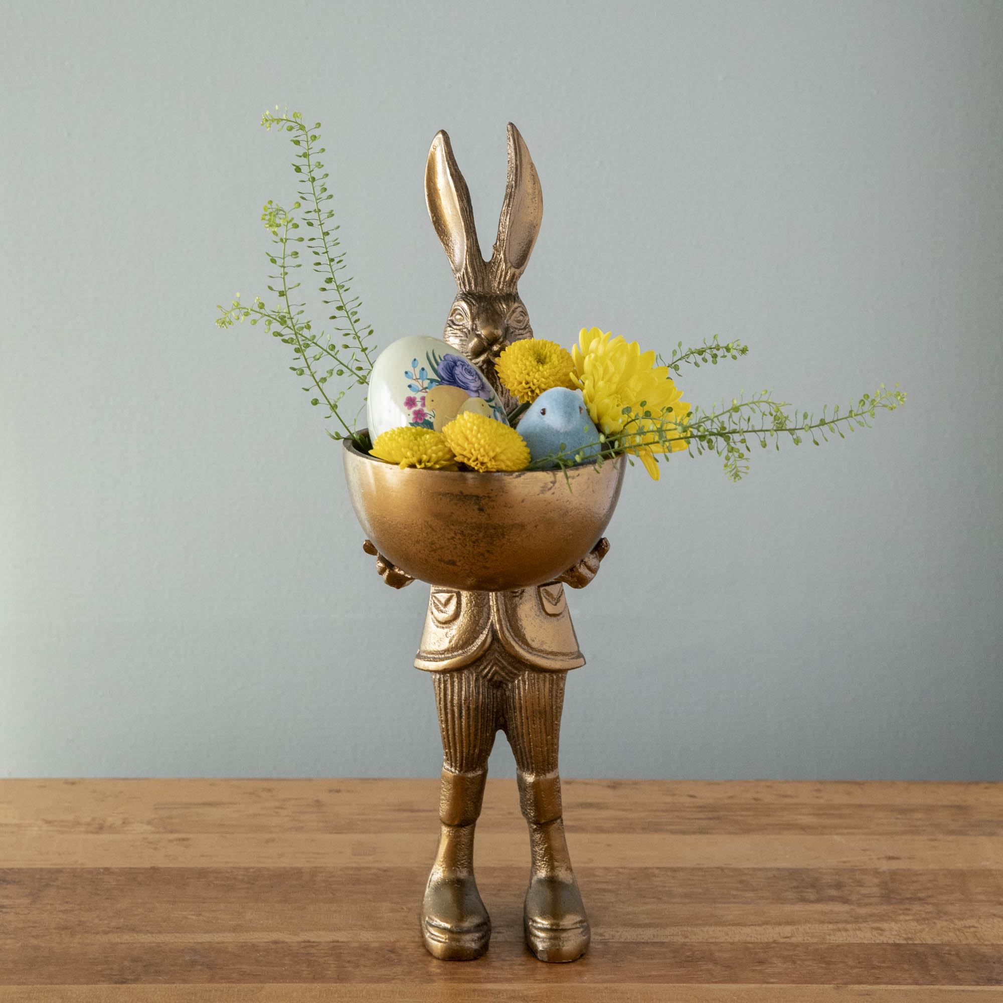 A well-dressed Hare Dish Stand by Accent Decor holding a round dish filled with a decorative egg, flocked chick and flowers. 