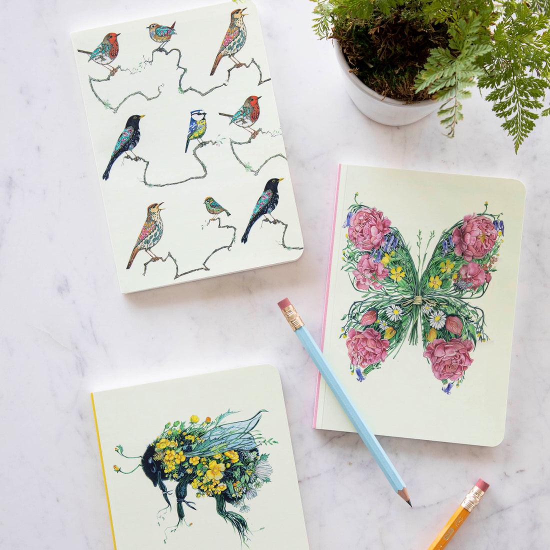 A Songbird Perfect Bound Notebook with a bird illustration on the cover surrounded by art supplies and white flowers on a green background, crafted by British papermakers on luxury cream stock, created by The DM Collection.