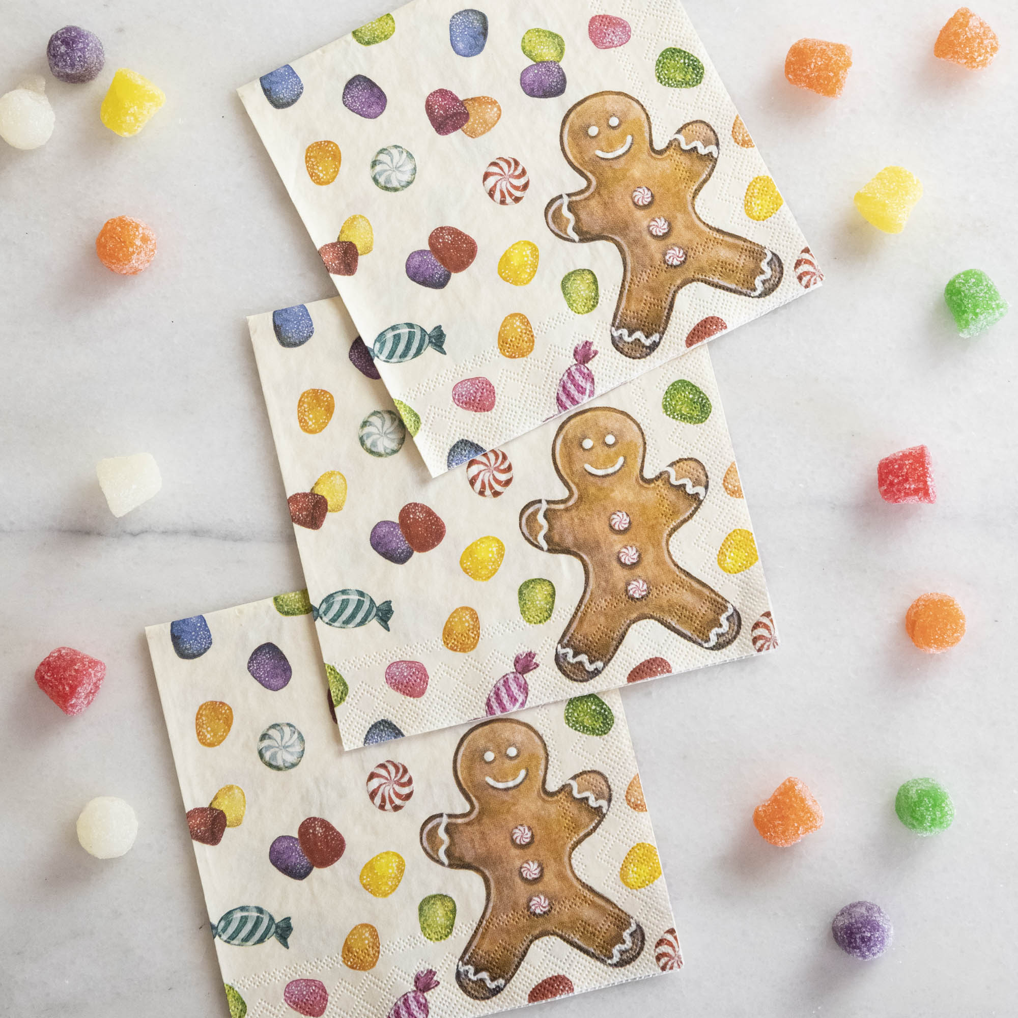 Three Gingerbread Cocktail Napkins overlapping on a white table surrounded by gumdrops.