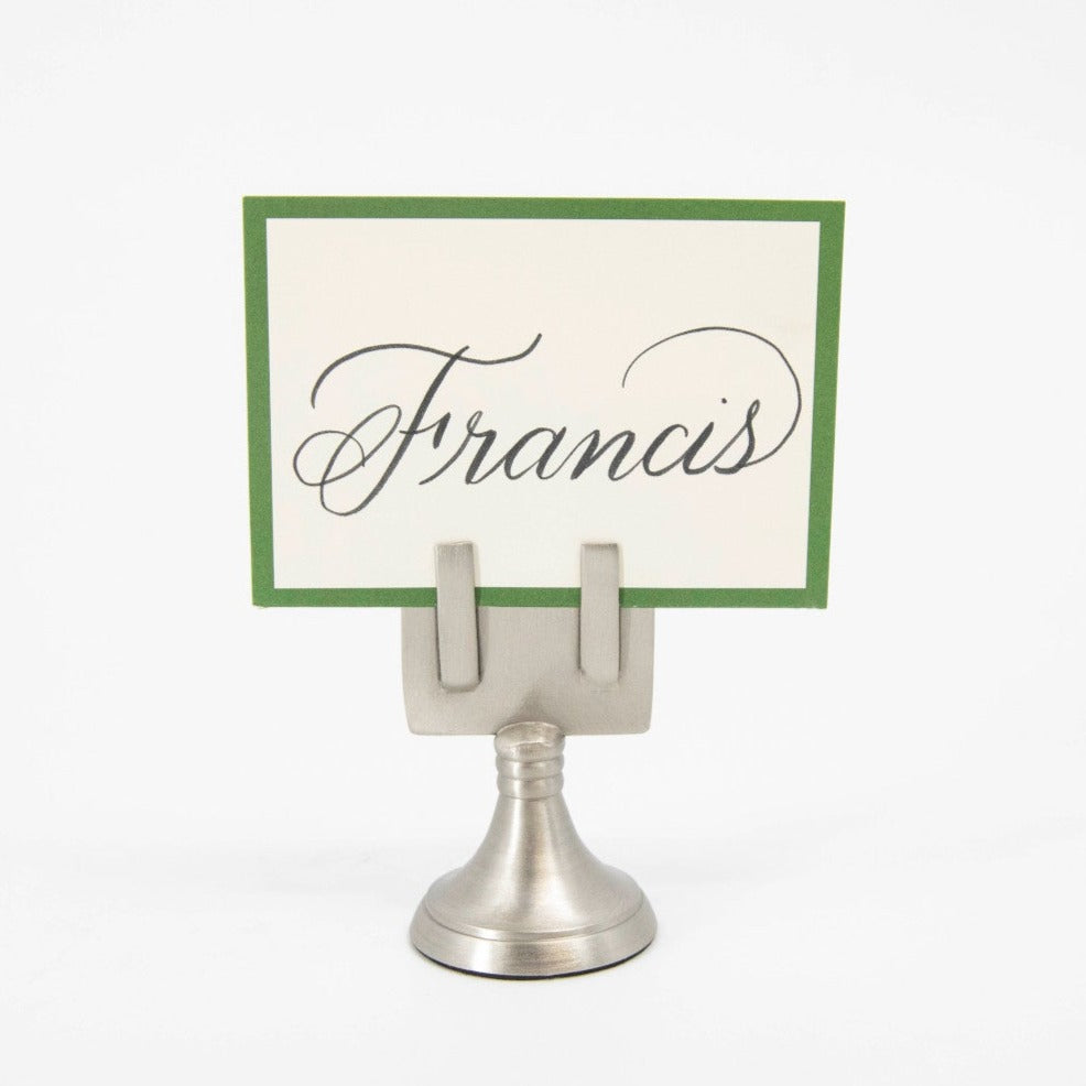 An elegant, silver, short round stand topped by a flat card-holder with two flat prongs, holding a place card reading &quot;Francis&quot;.